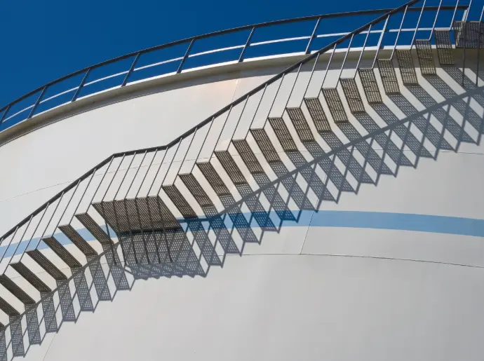 Industrial metal staircase against a white fuel storage tank under a clear sky / HSO Trading